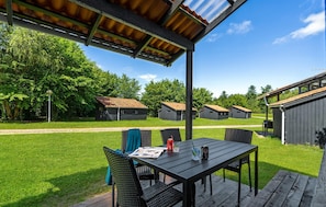 A covered outdoor seating area with a table and chairs, surrounded by a grassy lawn and small buildings in the background.