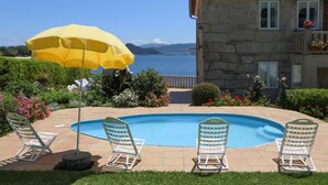 Zona de piscina con sillas de descanso y un paraguas amarillo, rodeada de jardines cuidados y un edificio de piedra con vistas al mar.