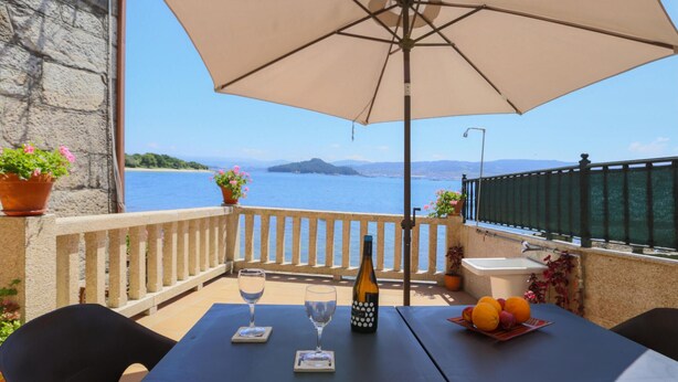 Una mesa con una botella de vino, una copa y fruta en una terraza con vista al agua.