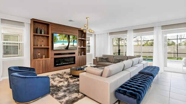 A modern living room with a large flat-screen TV, a wooden entertainment unit, a sofa, a blue ottoman, and a view of a pool through large windows. - 9BR house with game room near Disney area (Kissimmee)