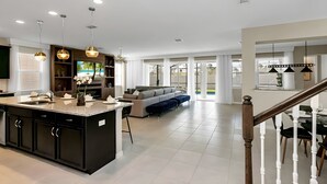 A modern kitchen with a central island, a dining area with a table and chairs, a living room with a sofa and a TV, and a staircase leading to another level. - 9BR house with game room near Disney area (Kissimmee)