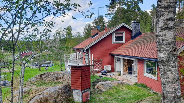 A red wooden house with a stone chimney, a porch, and a small garden area with rocks and trees. - 4 bedroom nice home in Trosa (Trosa)