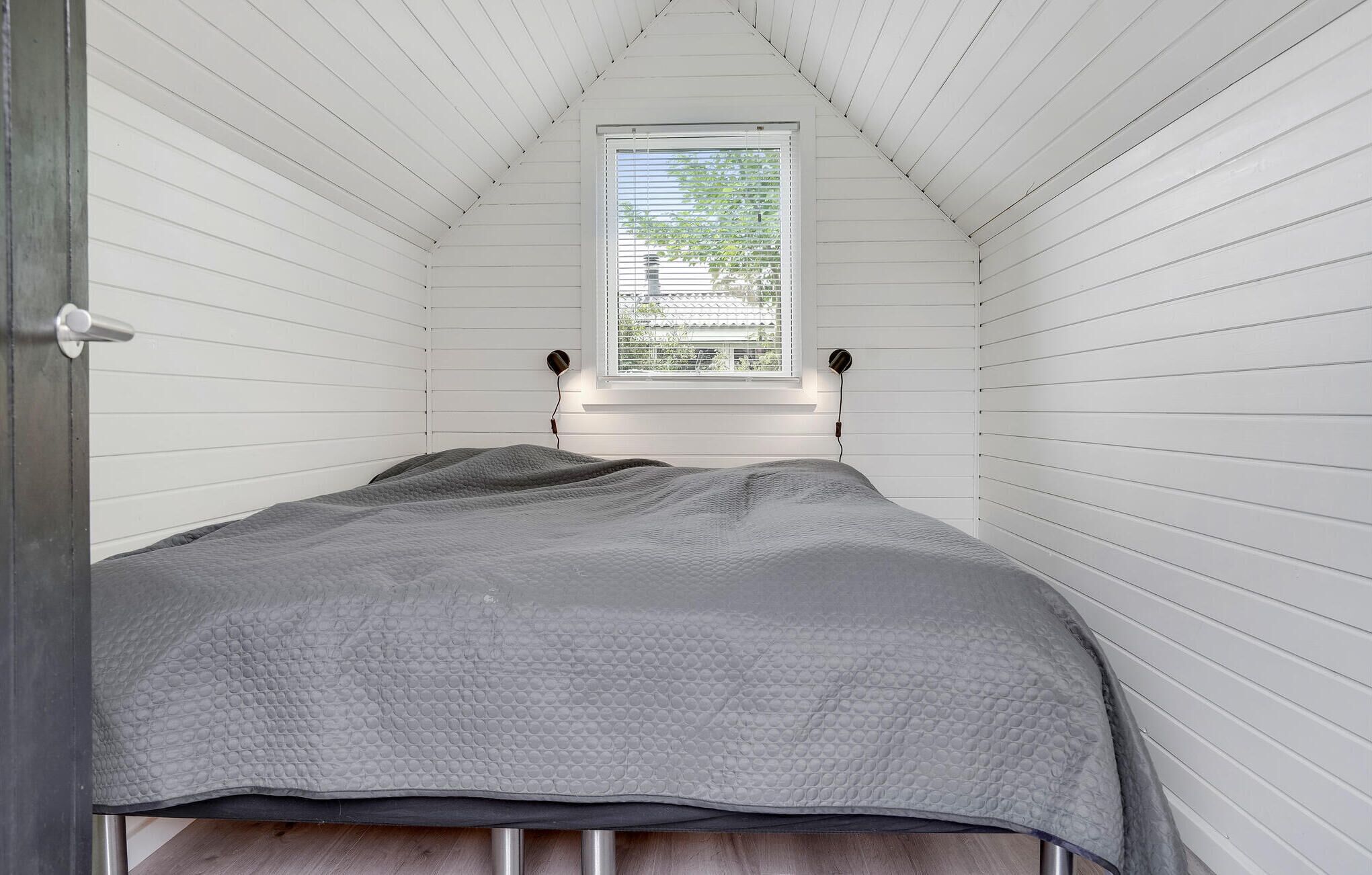 Une petite chambre avec un lit simple, aux murs en bois peints en blanc et au plafond en pente. On aperçoit une fenêtre avec des stores, et une lampe de chevet est fixée au mur.