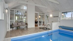 Een modern binnenzwembad met een helder zicht op het zwembad, een zithoek met een bank en tafel, en een afzonderlijke eethoek met stoelen.