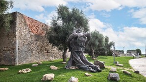 Une sculpture représentant une figure musclée dans une pose dynamique, entourée de rochers et d’herbe, avec un mur de pierre et des arbres en arrière-plan.