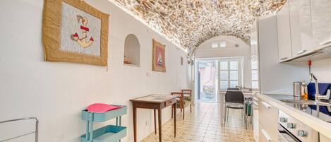 A kitchen with a tiled floor, a wooden table, chairs, a blue cart, and a hanging tapestry.