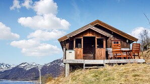 A wooden cabin with a porch and chairs, set against a mountainous backdrop.