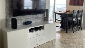 En moderne stue med et stort fladskærms-tv monteret på en trævæg, et hvidt tv-møbel med skuffer og et spiseområde med bord og stole.