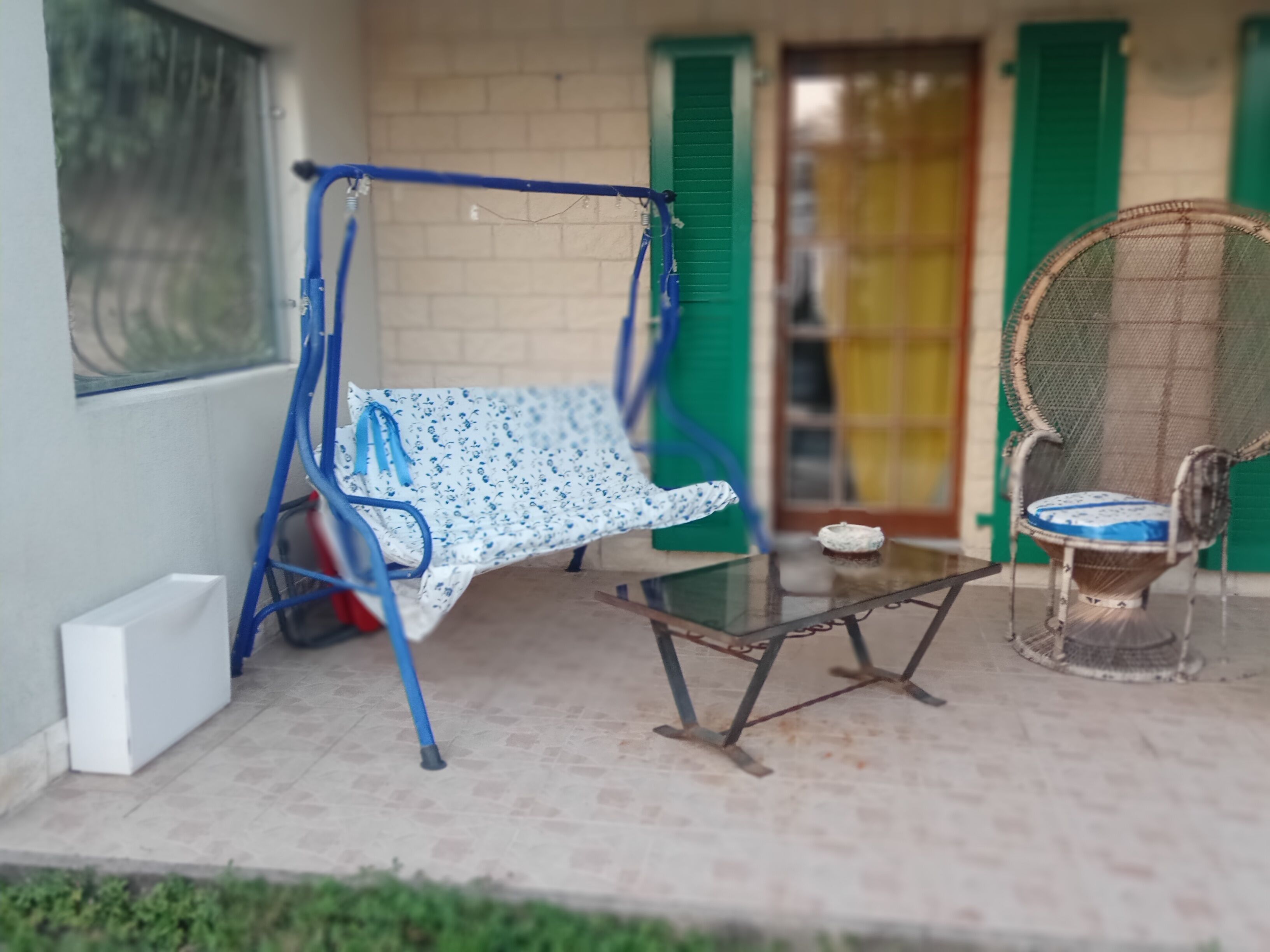 Een klein terras met een schommelstoel, een rieten stoel en een tafel.