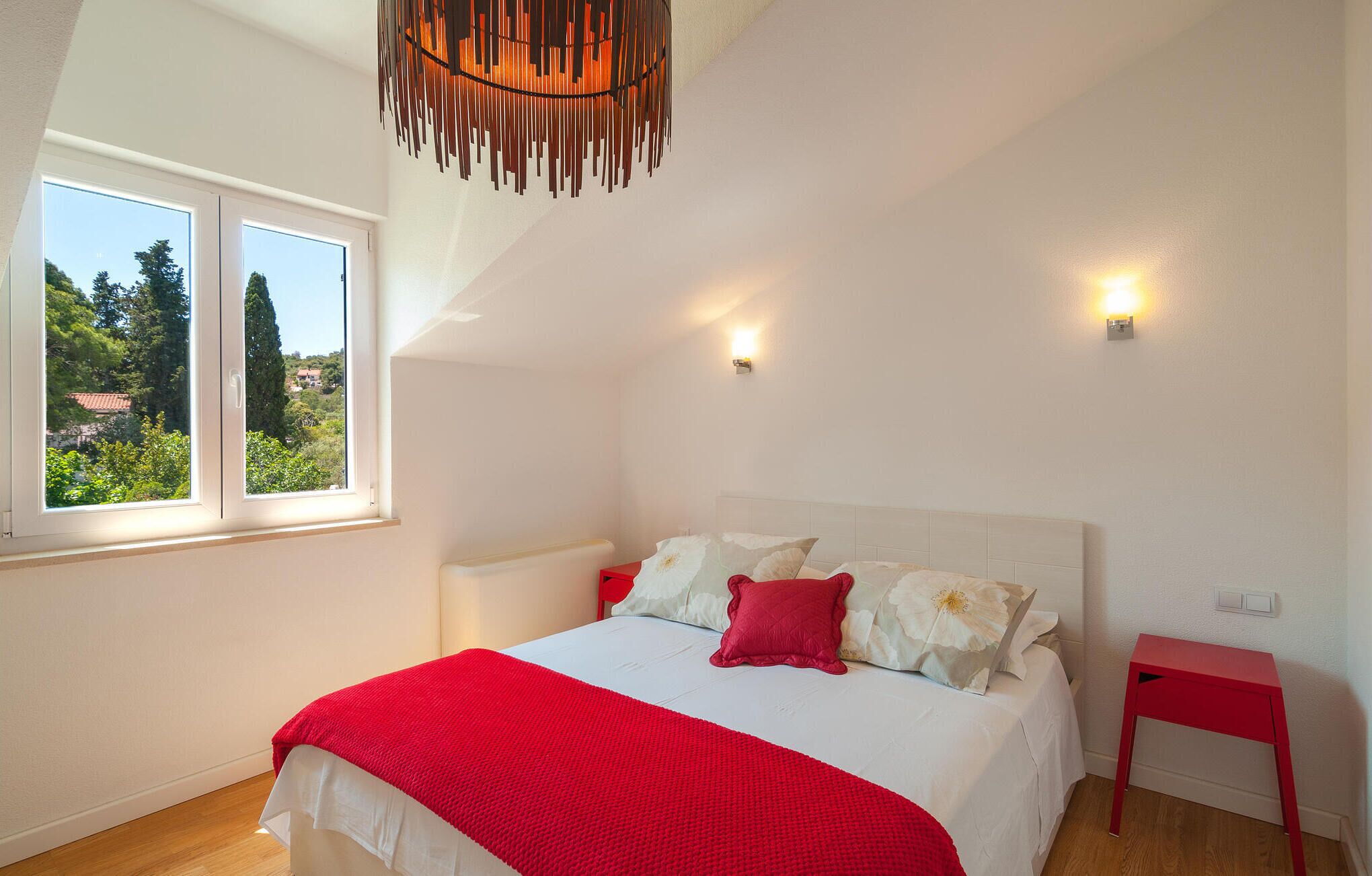 A bedroom with a bed, a window, a red bedside table, and a red chair.