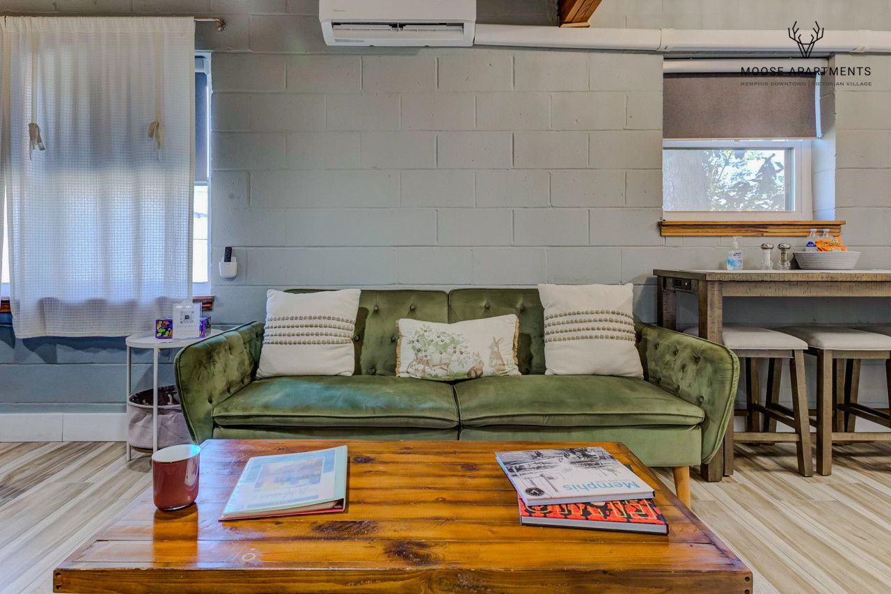 Un sofá verde con cojines blancos, una mesa de centro de madera con libros, una ventana con vistas y una zona de kitchenette.