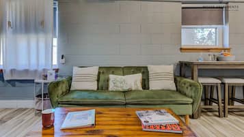 Un sofá verde con cojines blancos, una mesa de centro de madera con libros, una ventana con vistas y una zona de kitchenette.