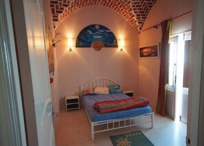 A bedroom with a bed, a nightstand, a wall-mounted lamp, a painting, and a window with curtains.