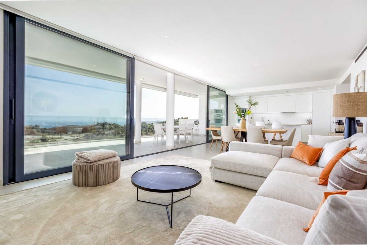 Un salon moderne doté d’une grande porte vitrée donnant sur un balcon, d’une table basse ronde, d’un canapé et d’un coin repas avec des chaises.