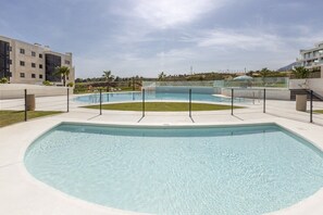 A swimming pool with a surrounding fence, a grassy area, and modern buildings in the background.