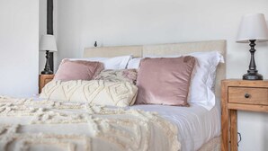 A neatly made bed with a beige and pink pillow arrangement, a wooden nightstand with a lamp, and a plain white wall. - Stunning Character 2bed Cottage in St Albans Wifi (St Albans)