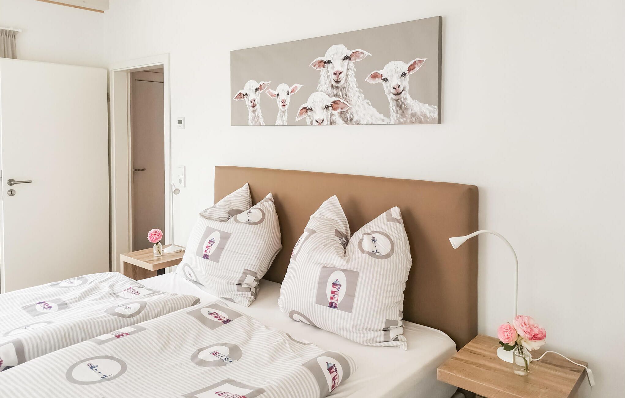 A hotel room with two beds, a headboard, bedside tables, a lamp, and a painting of sheep on the wall.