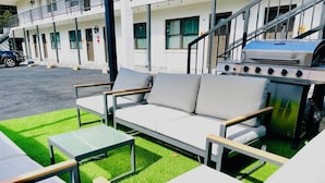 Et moderne udendørs opholdsområde med en sofa og borde, grill og en bygning med balkoner i baggrunden.
