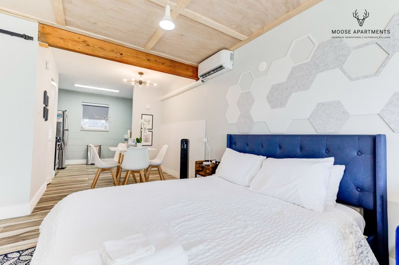 A bedroom with a large bed, a blue headboard, a desk, and a chair. There is a window with blinds, a wall-mounted air conditioner, and a ceiling fan.