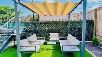 Un área de estar al aire libre cubierta con sillas blancas y una mesa, rodeada de césped artificial verde y un seto.