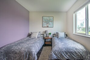 A bedroom with two single beds, a nightstand with a lamp, a small table, and a window with a view of the outside. - Entire home remodeled & 1 mile to Kenai River & 1/2 block to Peninsula Hospital (Soldotna)