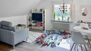 Ein Wohnzimmer mit grauem Sofa, einem Fernseher, einem Bücherregal und einem Spielbereich mit Spielzeugautos.