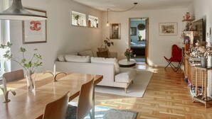 Ein modernes Wohnzimmer mit Sofa, Couchtisch und Essbereich mit Stühlen und Tisch. Ein Bücherregal mit Dekorationselementen und ein Spiegel sind ebenfalls vorhanden.
