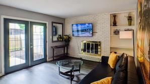Ein Wohnzimmer mit einer schwarzen Ledersofa, einem Glastisch, einem Kamin und einem an einer Ziegelwand befestigten Fernseher.