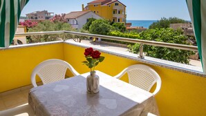 A balcony with a yellow wall, white chairs, a table with a vase of red roses, and a view of the sea and buildings.