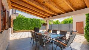 Un espace extérieur abrité, aménagé pour prendre les repas, avec une table et des chaises, une plante suspendue, et une vue sur une haie verdoyante et un bâtiment en arrière-plan.