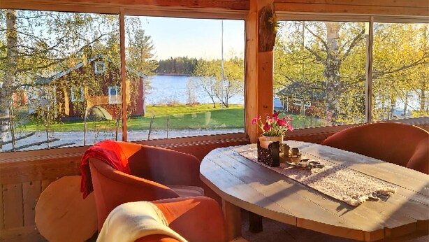 A cozy room with a wooden table and chairs, large windows with a view of a lake and greenery.