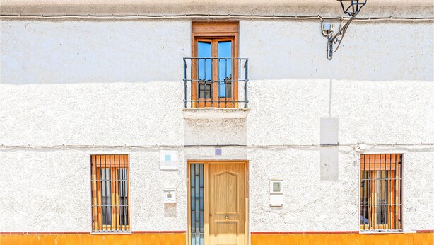 Un edificio con puerta de madera, dos ventanas con rejas de metal y un balcón con barandilla metálica.