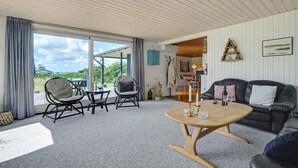 Ett rymligt vardagsrum med ett stort fönster, ett träpanelat tak, en svart lädersoffa och ett runt träbord.