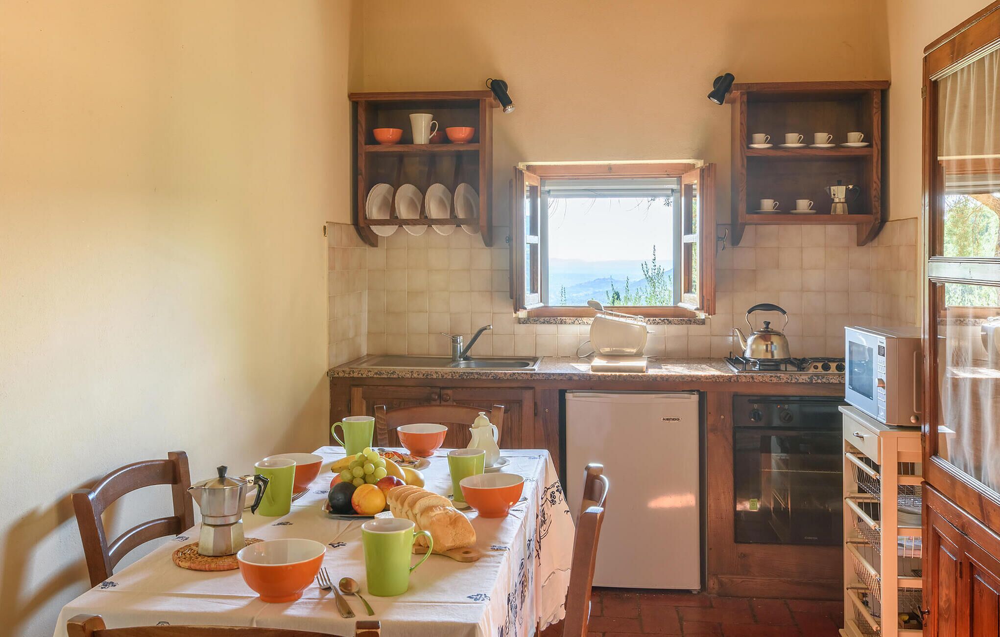 Une cuisine compacte avec une table à manger préparée pour le petit-déjeuner, un réfrigérateur et une fenêtre donnant sur un beau paysage.