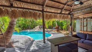 A pool area with a thatched roof, wicker furniture, and palm trees.