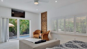 A modern bedroom with a large bed, a sofa, a dining table with chairs, and a view of a tropical garden through the sliding glass doors.