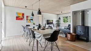 Moderni ruokailutila, jossa on pitkä pöytä, mustat tuolit ja riippuvalaisimet. Seinällä on televisio ja suuri ikkuna, josta avautuu näkymä puita kohti.