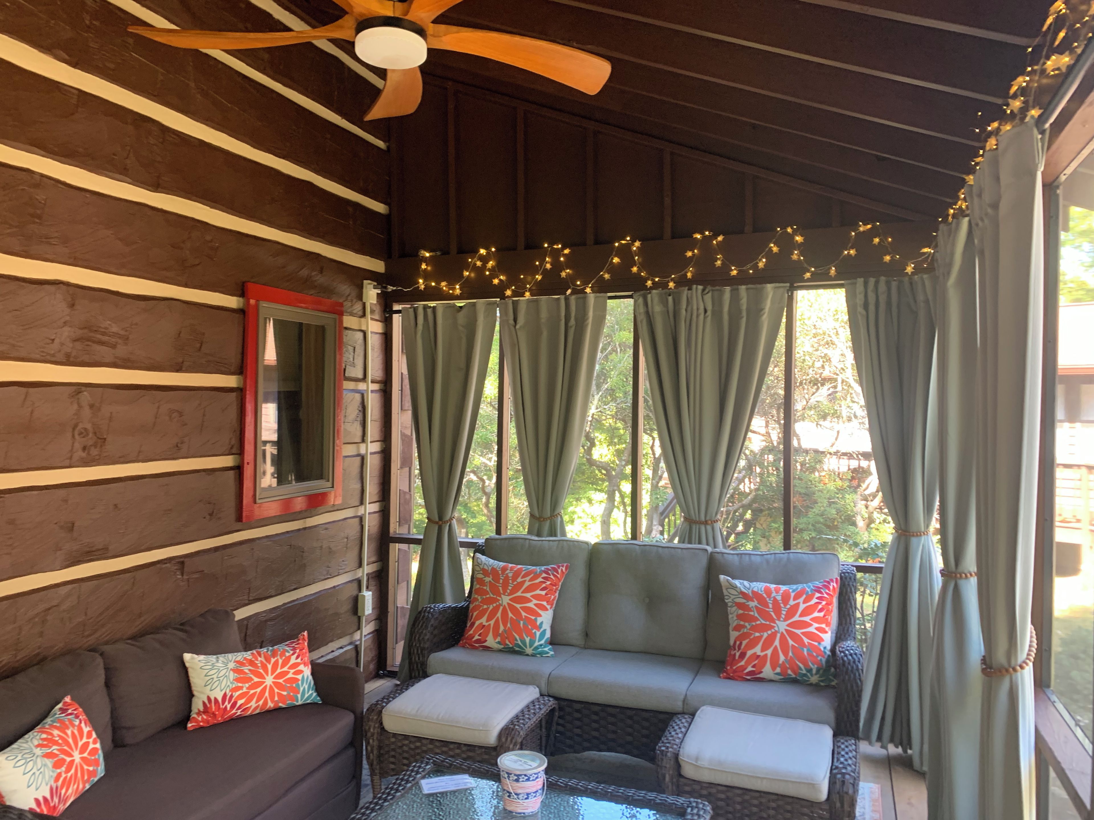A cozy outdoor seating area with a ceiling fan, string lights, and a view of greenery outside.