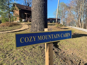 A sign for 'Cozy Mountain Cabin' in a wooded area with a cabin in the background.