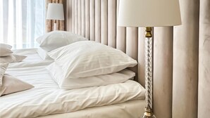 A neatly made bed with white linens and pillows, a bedside lamp, and a curtain.