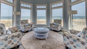 Una habitación con amplios ventanales con vistas a una playa, con muebles de estampado floral y un otoman central.