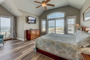 Un dormitorio con una cama grande, un ventilador de techo, un aparador y un televisor. Hay ventanas amplias con vista a la playa.