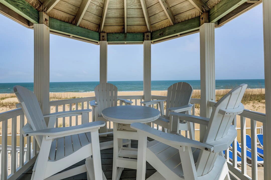 A covered outdoor seating area with white chairs and a round table, overlooking a beach.
