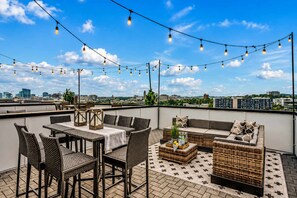 Un espace extérieur aménagé avec des meubles en osier, une table et des guirlandes lumineuses, le tout sur fond de panorama urbain.