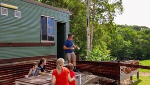 A rustic cabin with a green exterior, a wooden deck, and a picnic table where a family is seated. - Kur Cow Farmstay (Kuranda)