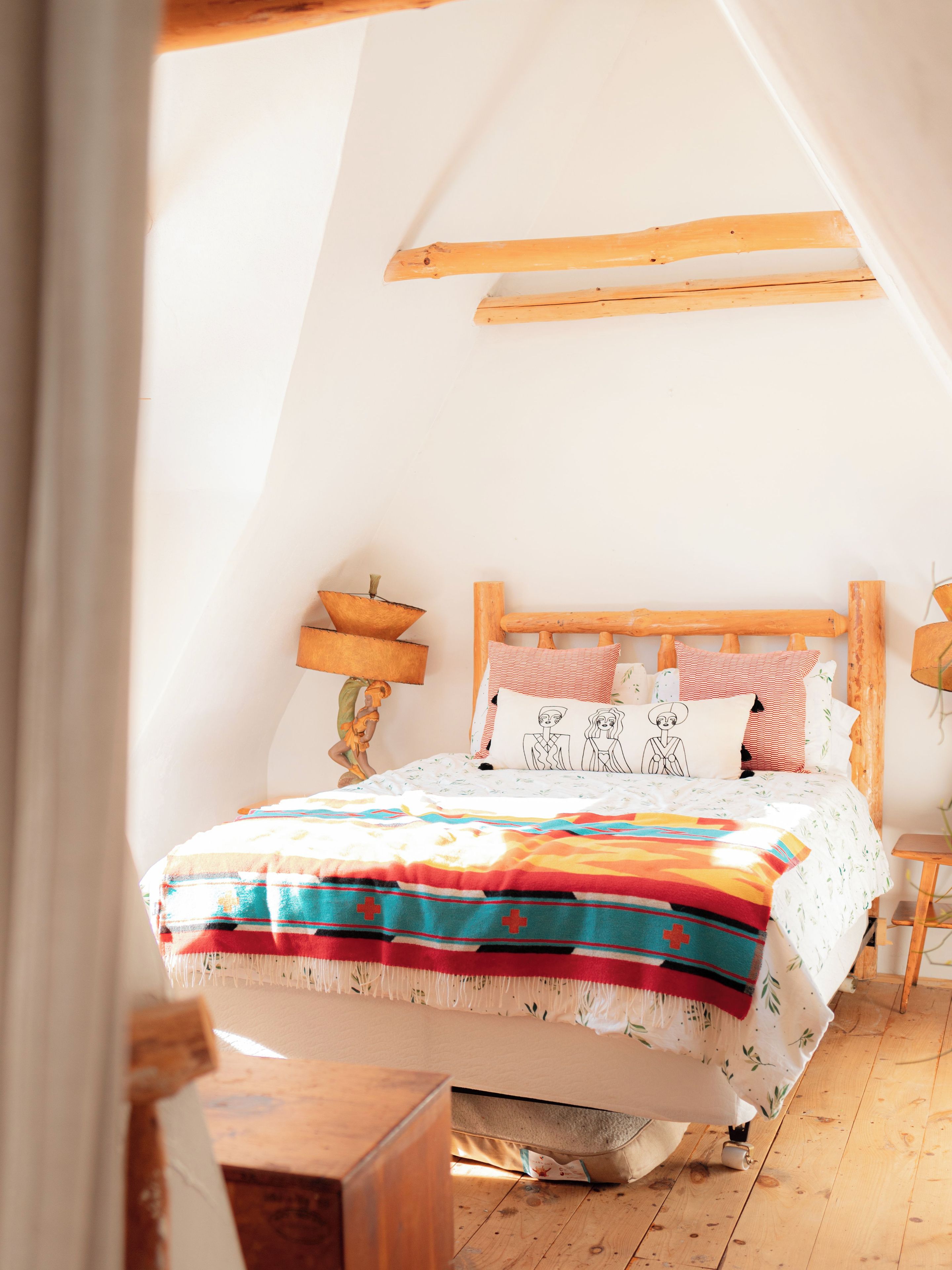 Une chambre avec un plafond en pente, un cadre de lit en bois, une courtepointe aux couleurs vives et une table de chevet avec une lampe.