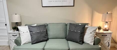A green sofa with three pillows, a framed sign above, a lamp on a bedside table, and a small plant.