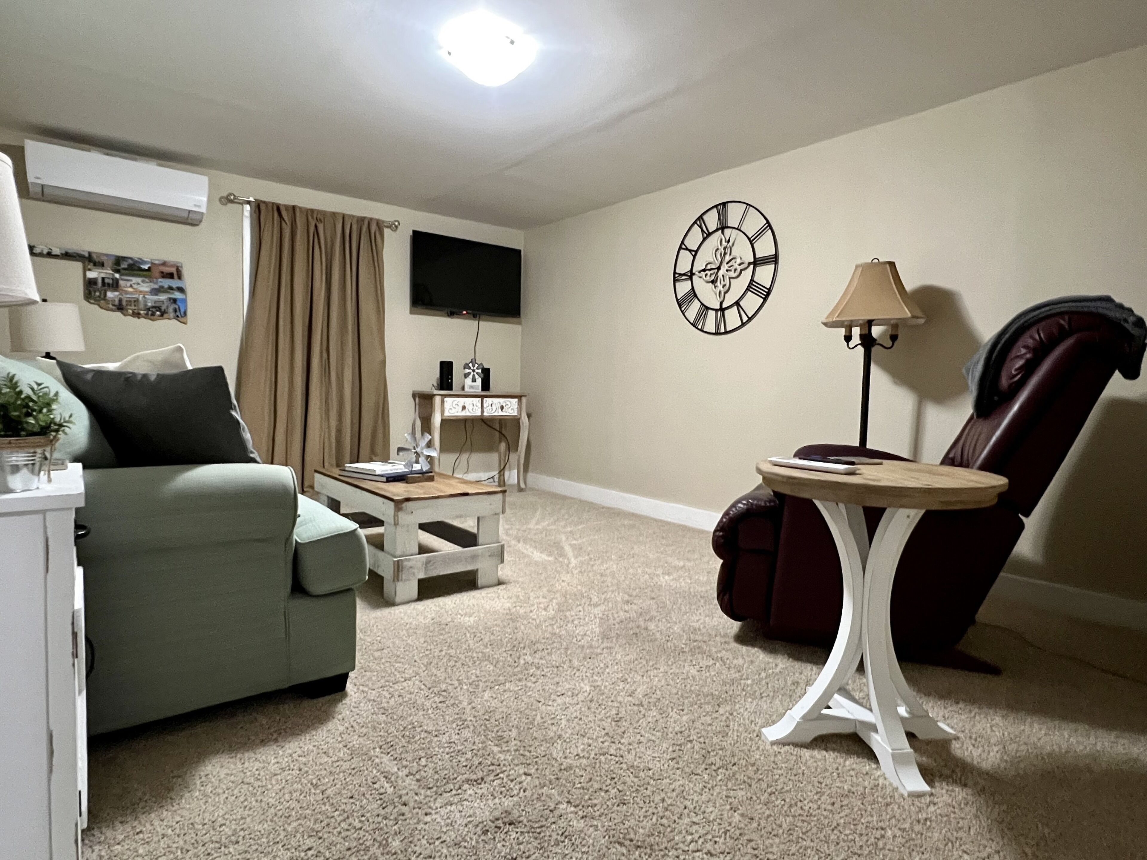 A living room with a sofa, a small table, a chair, a clock on the wall, and a television.
