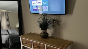 En stue med et ophængt fjernsyn der viser streamingtjenester, et træsofabord med en vase, og et spejl der reflekterer en seng og gardiner.