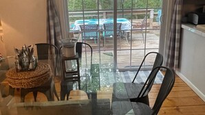 Un coin repas avec une table et des chaises en verre, un espace cuisine avec des placards, et une vue sur une terrasse extérieure équipée d’une table et de chaises.